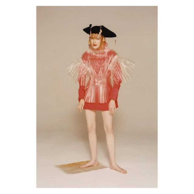 A person poses in avant-garde fashion featuring a red dress, a large brimmed hat, and a translucent vest-like piece in a studio setting.