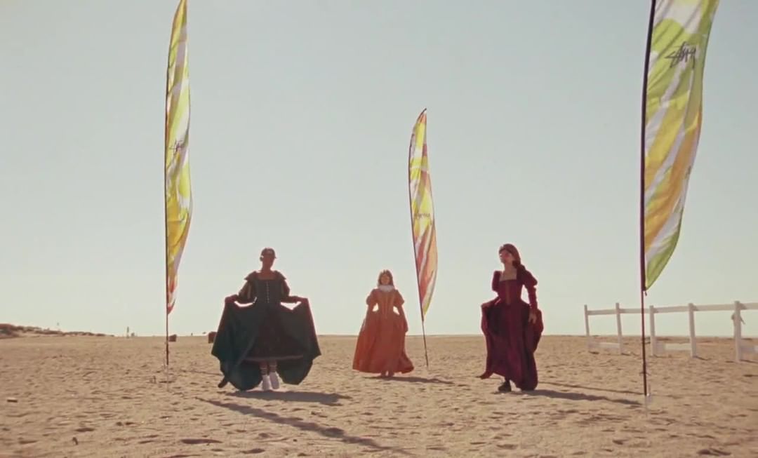 Three women in period dresses walk on a sunny beach with colorful advertising flags in the background, creating a blend of historical and modern.