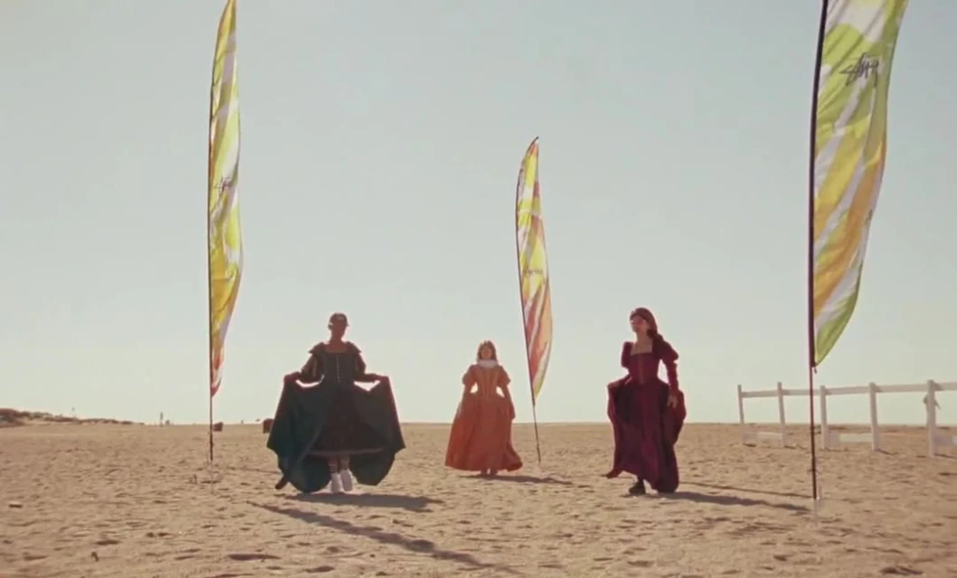 Three women in period dresses walk on a sunny beach with colorful advertising flags in the background, creating a blend of historical and modern.