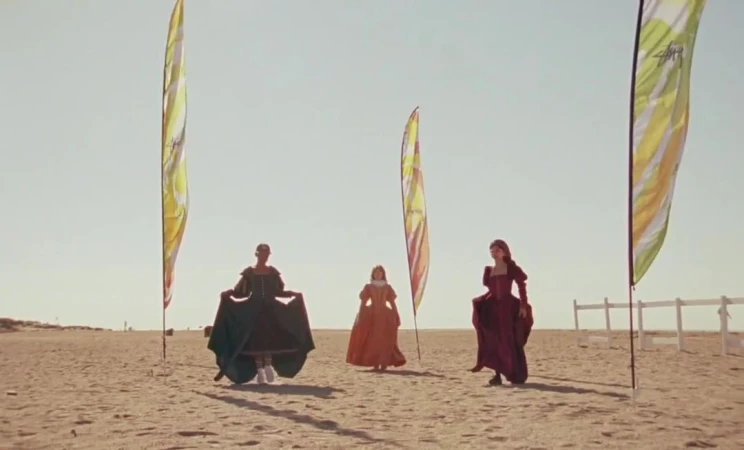 Three women in period dresses walk on a sunny beach with colorful advertising flags in the background, creating a blend of historical and modern.