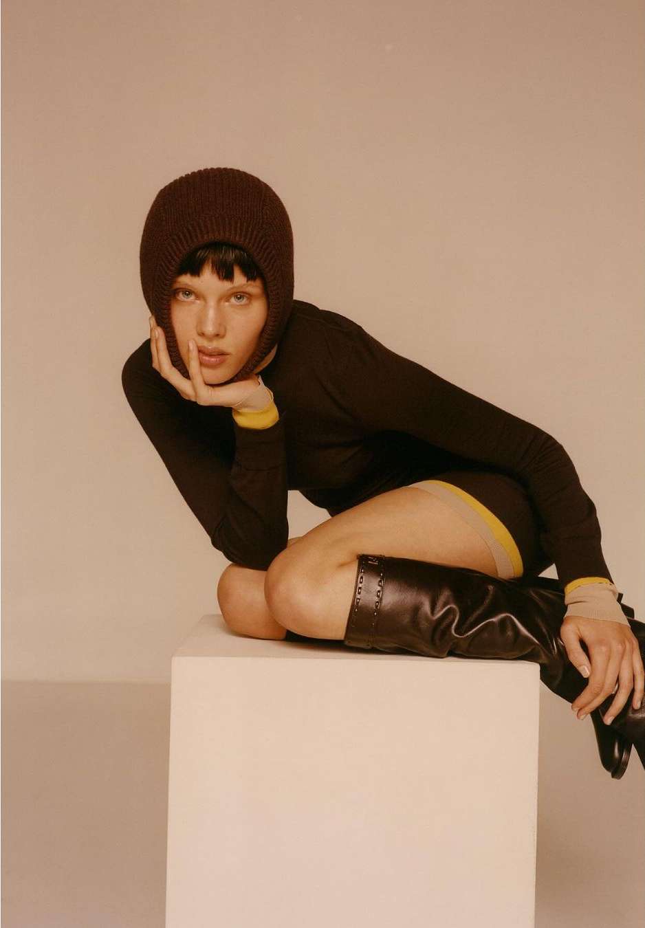 A woman poses in a studio wearing a brown knit balaclava and long-sleeved shirt, with black boots on a white cube in a studio setting.