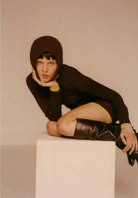 A woman poses in a studio wearing a brown knit balaclava and long-sleeved shirt, with black boots on a white cube in a studio setting.