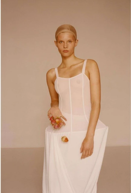 A fair-skinned woman with a nude cap poses with fruit, wearing a white bodice and dress against a neutral background in a studio shot.