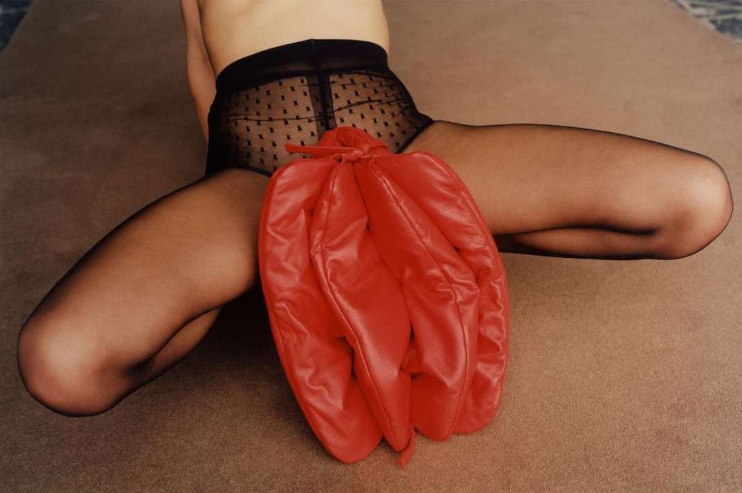 Seated person in black patterned tights with a bright red leather drawstring bag between their legs, against a neutral background.