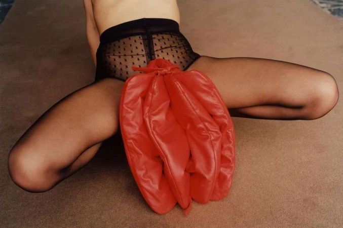 Seated person in black patterned tights with a bright red leather drawstring bag between their legs, against a neutral background.