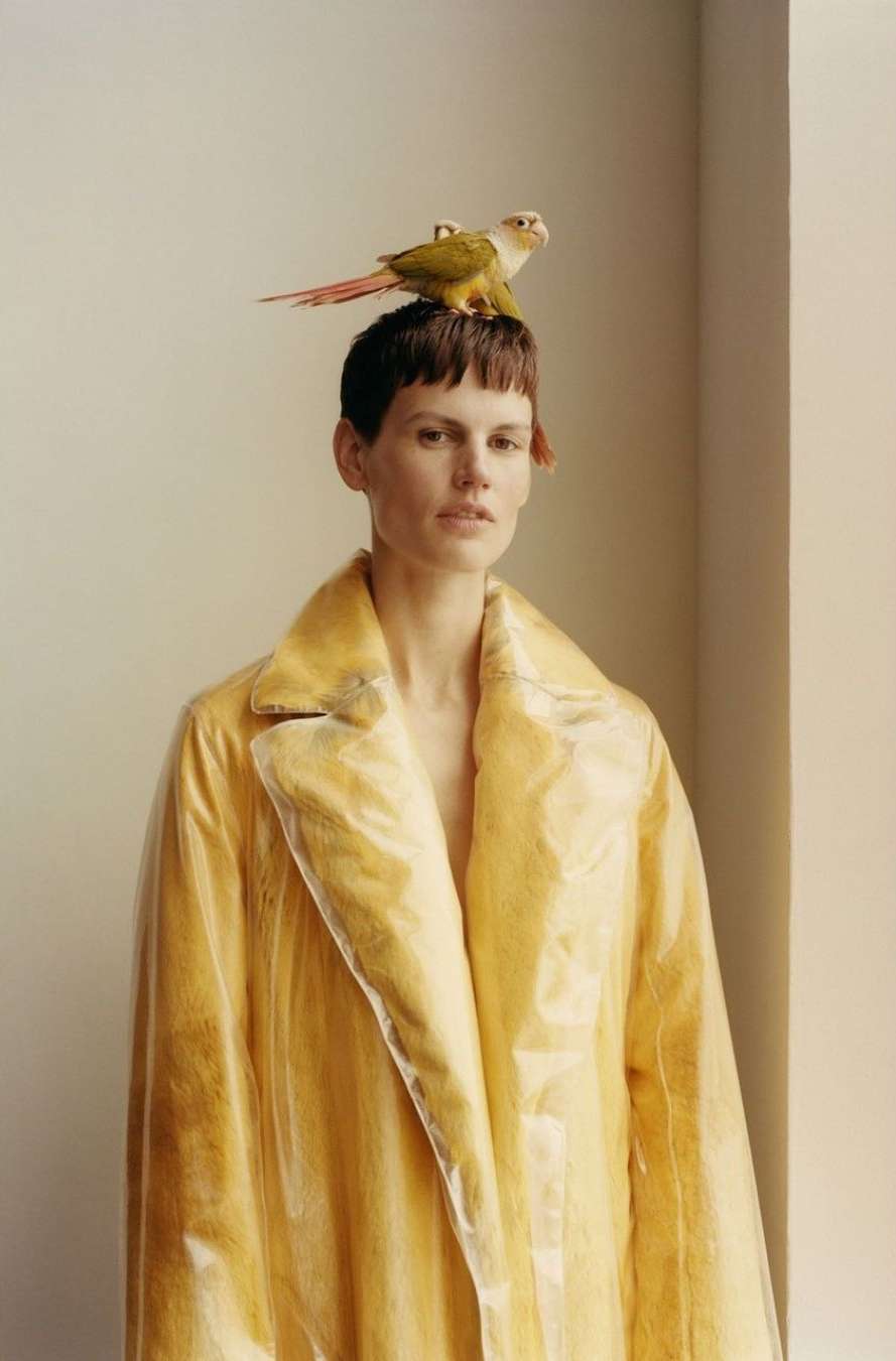 Adult woman wearing a yellow transparent trench coat poses indoors with a green and orange conure bird perched on her head.