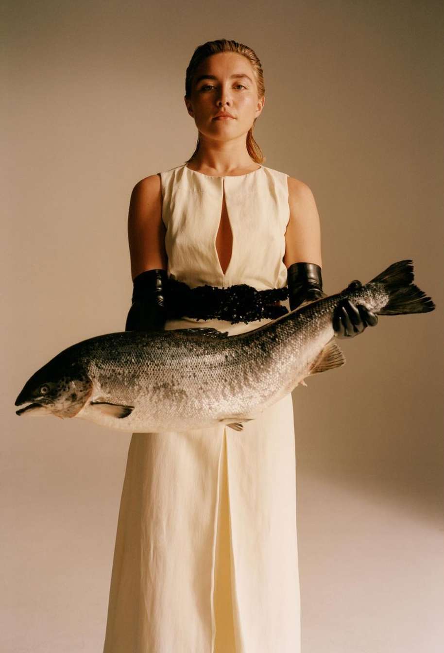Woman in dress holding a large fish in studio setting, featuring editorial style, neutral tones, and a surreal juxtaposition.