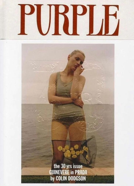 Fashion editorial photo features a female model in a tank top, briefs, and sheer skirt with yellow flowers, posing on a beach.