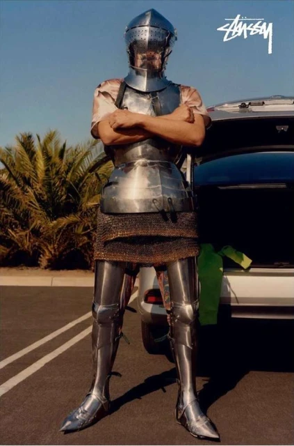 A person in full medieval armor stands in front of a car in a parking lot with palm trees and a blue sky in the background.