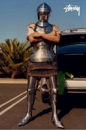 A person in full medieval armor stands in front of a car in a parking lot with palm trees and a blue sky in the background.