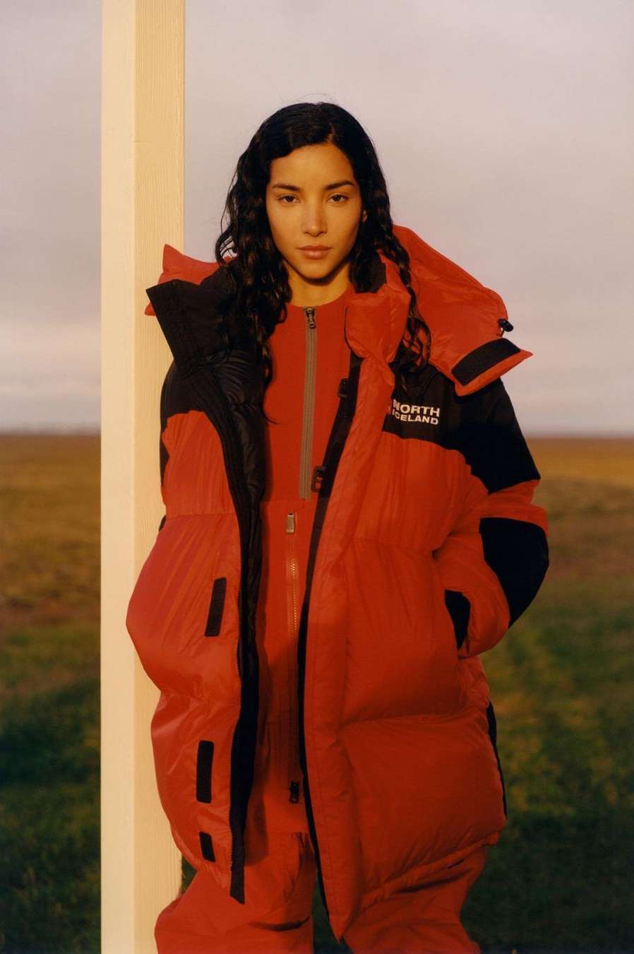 Woman in red puffer jacket and matching jumpsuit poses against a white post in a field, outdoors with a clear horizon backdrop.