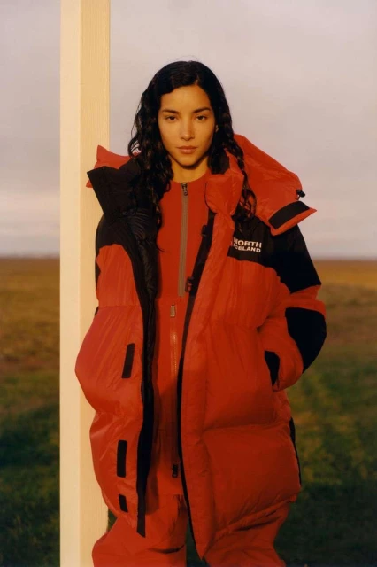 Woman in red puffer jacket and matching jumpsuit poses against a white post in a field, outdoors with a clear horizon backdrop.