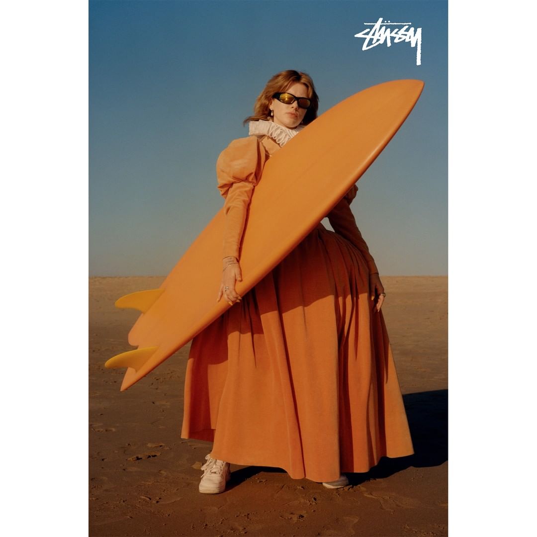 Stylish woman in orange dress poses with surfboard on sandy beach under clear sky; Stussy logo visible; fashion and sport.