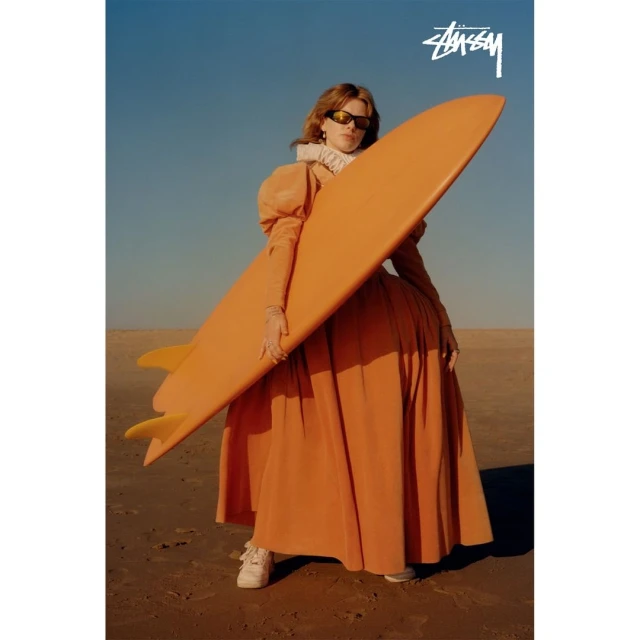 Stylish woman in orange dress poses with surfboard on sandy beach under clear sky; Stussy logo visible; fashion and sport.