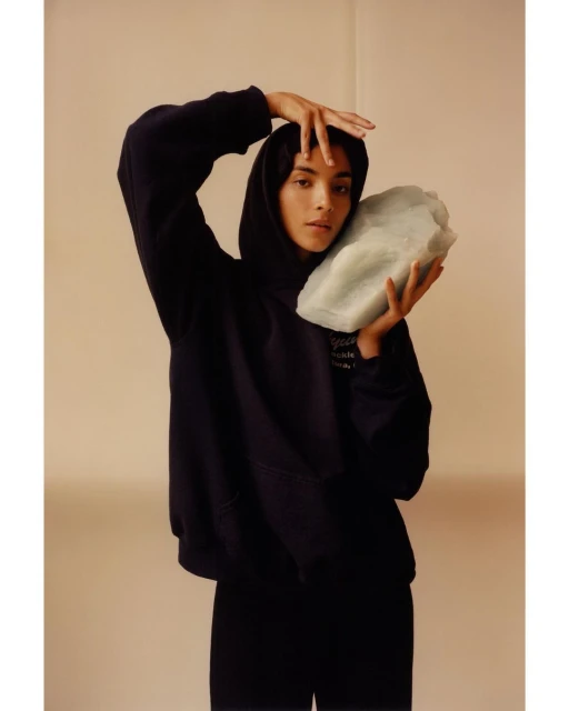 Portrait of woman in hoodie holding light green rock in front of neutral background in a fashion or beauty context.