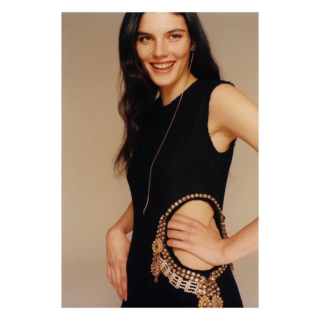 Smiling woman in a black dress with embellished cut-out and long gold earring posing in a studio with a light beige background.