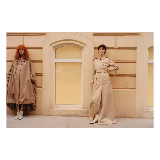 Two fashion models pose outdoors in oversized trench coats against a tan wall with an arched window in a fashion editorial.
