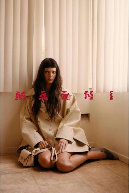 Woman in beige coat and grey shoes poses indoors sitting on floor with small purse and pink Marni logo in front of off-white curtains.