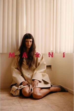 Woman in beige coat and grey shoes poses indoors sitting on floor with small purse and pink Marni logo in front of off-white curtains.
