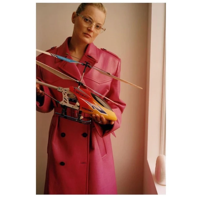 Adult female in pink coat holds an orange-and-yellow toy helicopter in a neutral indoor setting with a cream-colored wall and window.