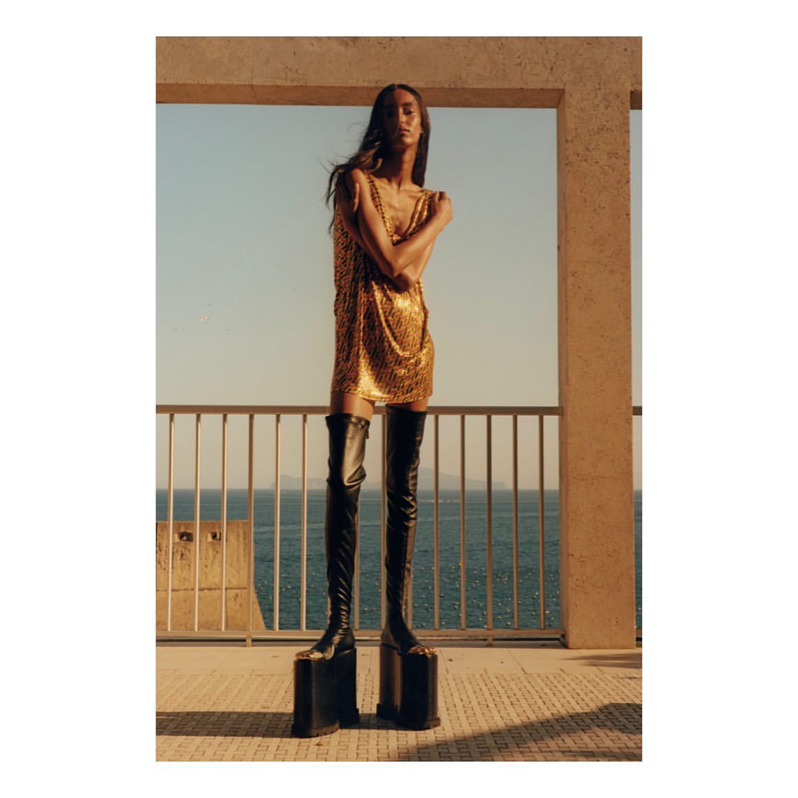 Fashion shot of a slender female model in a gold mini dress and black platform boots, posing on a balcony with a sea view.
