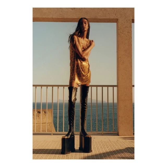Fashion shot of a slender female model in a gold mini dress and black platform boots, posing on a balcony with a sea view.