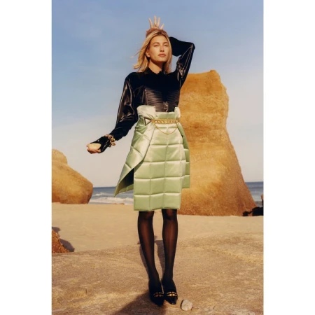 Model in black shiny blouse and quilted sage green skirt standing outdoors on a sandy beach with rock formations.