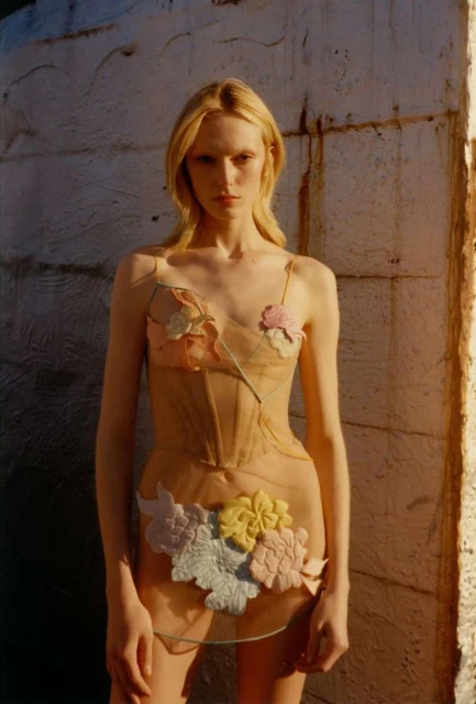 A model poses in a sheer beige dress adorned with pastel floral appliques, standing against a textured wall in soft, natural lighting.