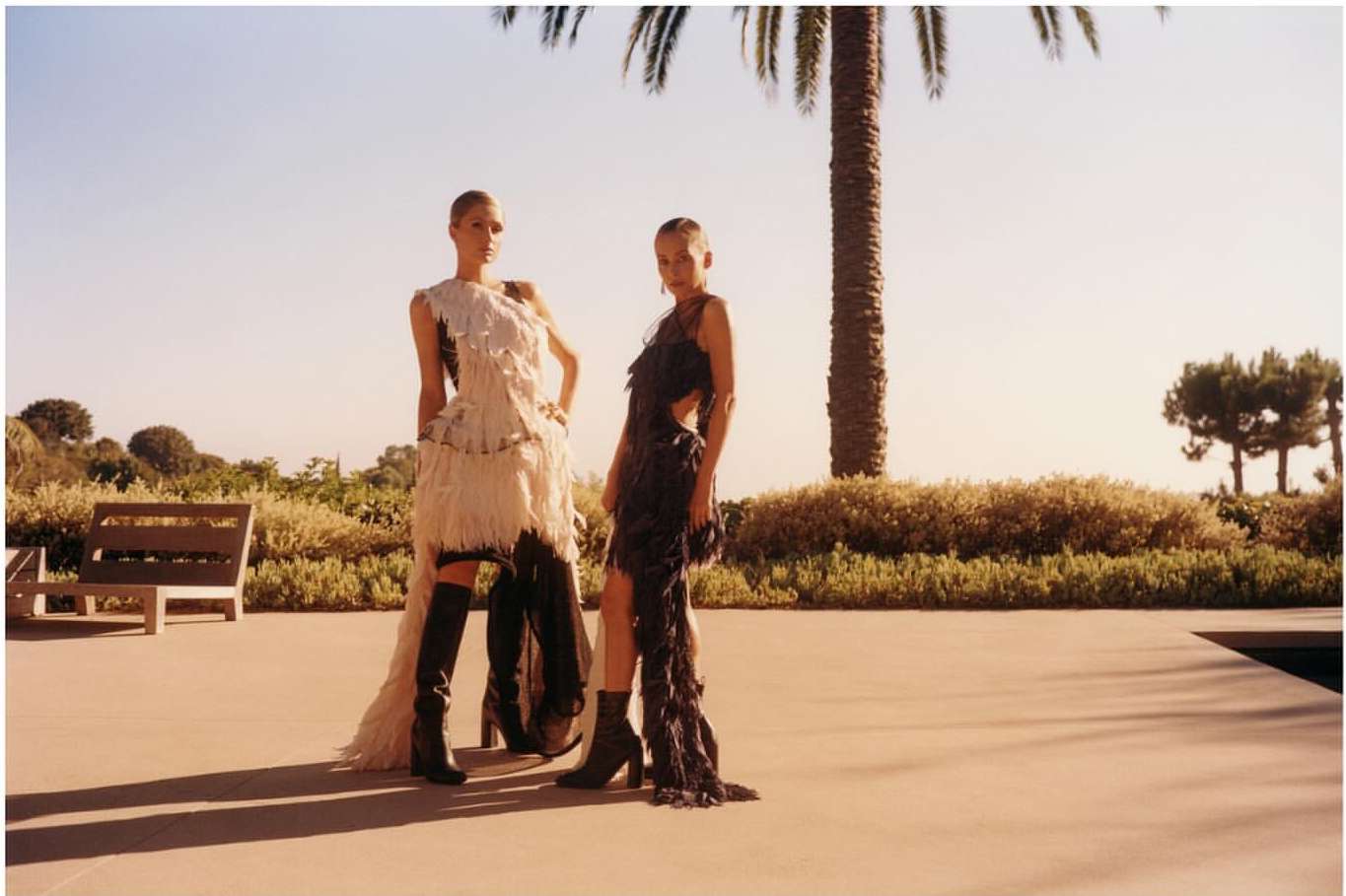 Two fashion models pose outdoors in feather dresses and boots with a palm tree and clear blue sky in the background on a sunny day.