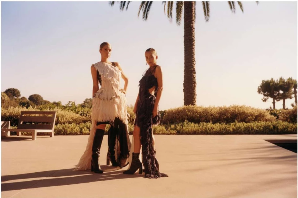 Two fashion models pose outdoors in feather dresses and boots with a palm tree and clear blue sky in the background on a sunny day.