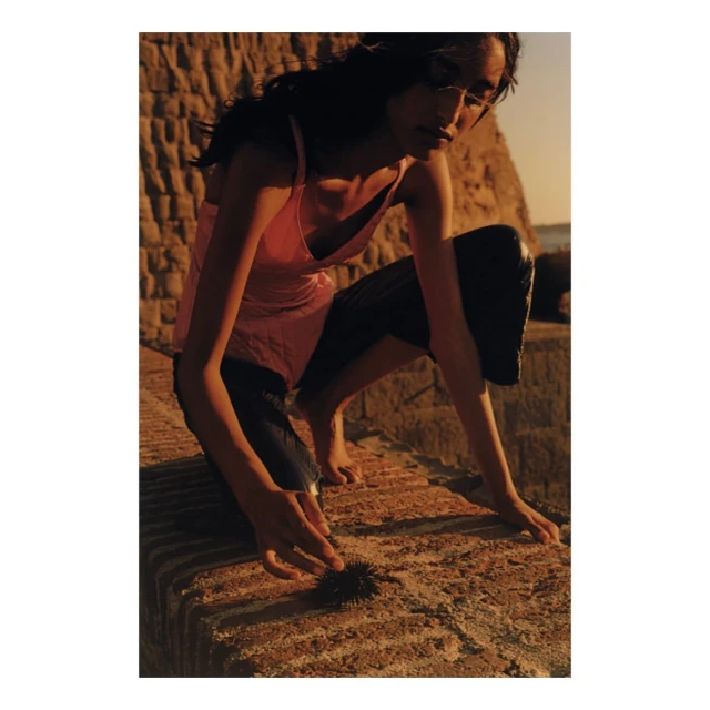 Woman in pink tank top crouches outdoors on brick surface, examining a dark spiky object with sunlight, ocean, and sky visible.
