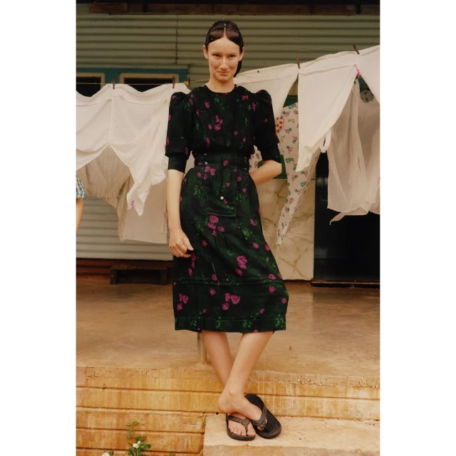 A young adult female standing on a porch, wearing a dark floral dress and sandals, with a clothesline in the background on a sunny day.