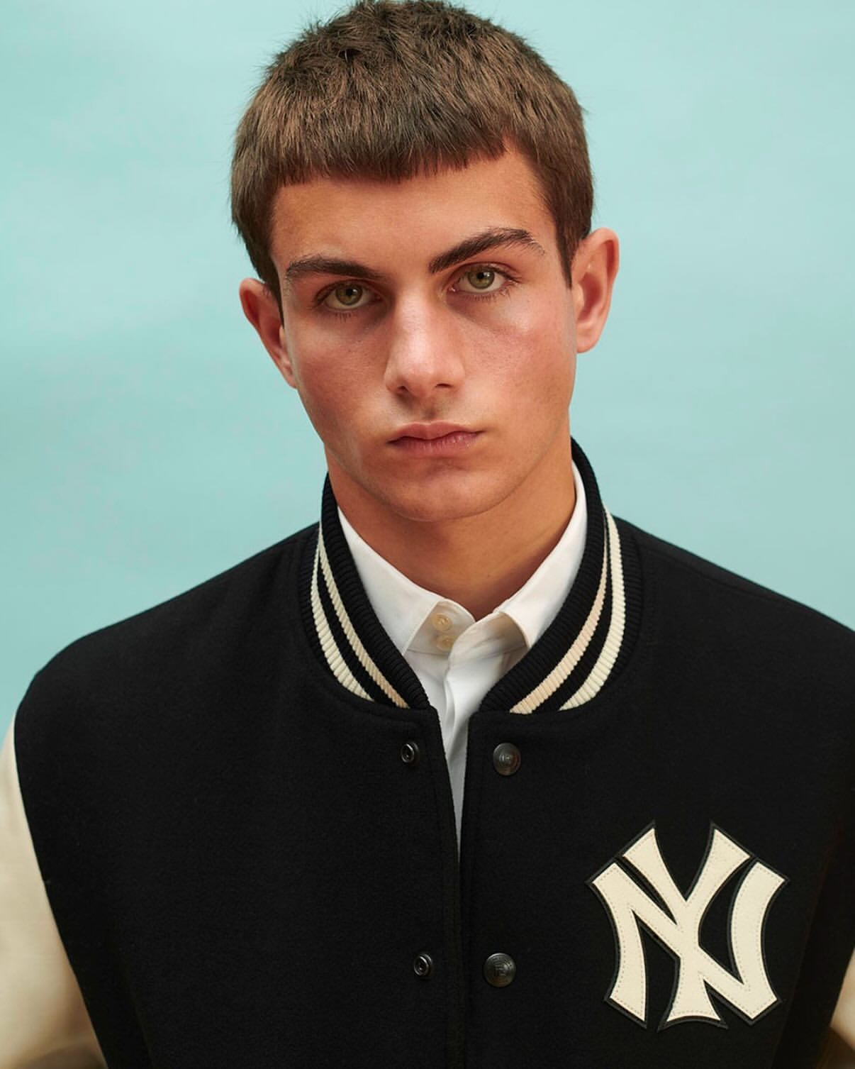 Close-up studio shot of a young adult male wearing a letterman jacket with an NY logo against a light blue background.