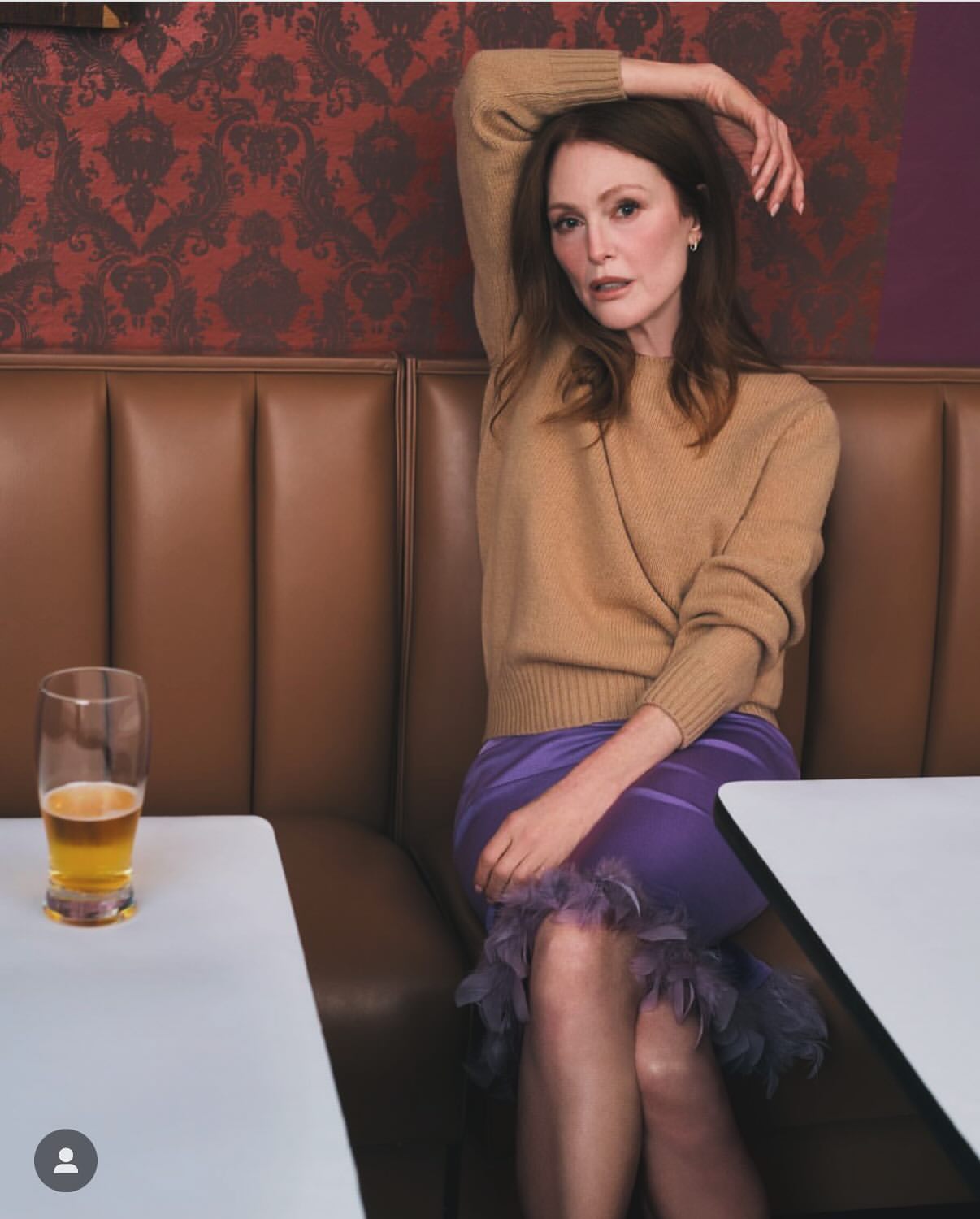 Indoor portrait of an adult woman with auburn hair, wearing a tan sweater and lavender skirt, seated at a table with a drink.