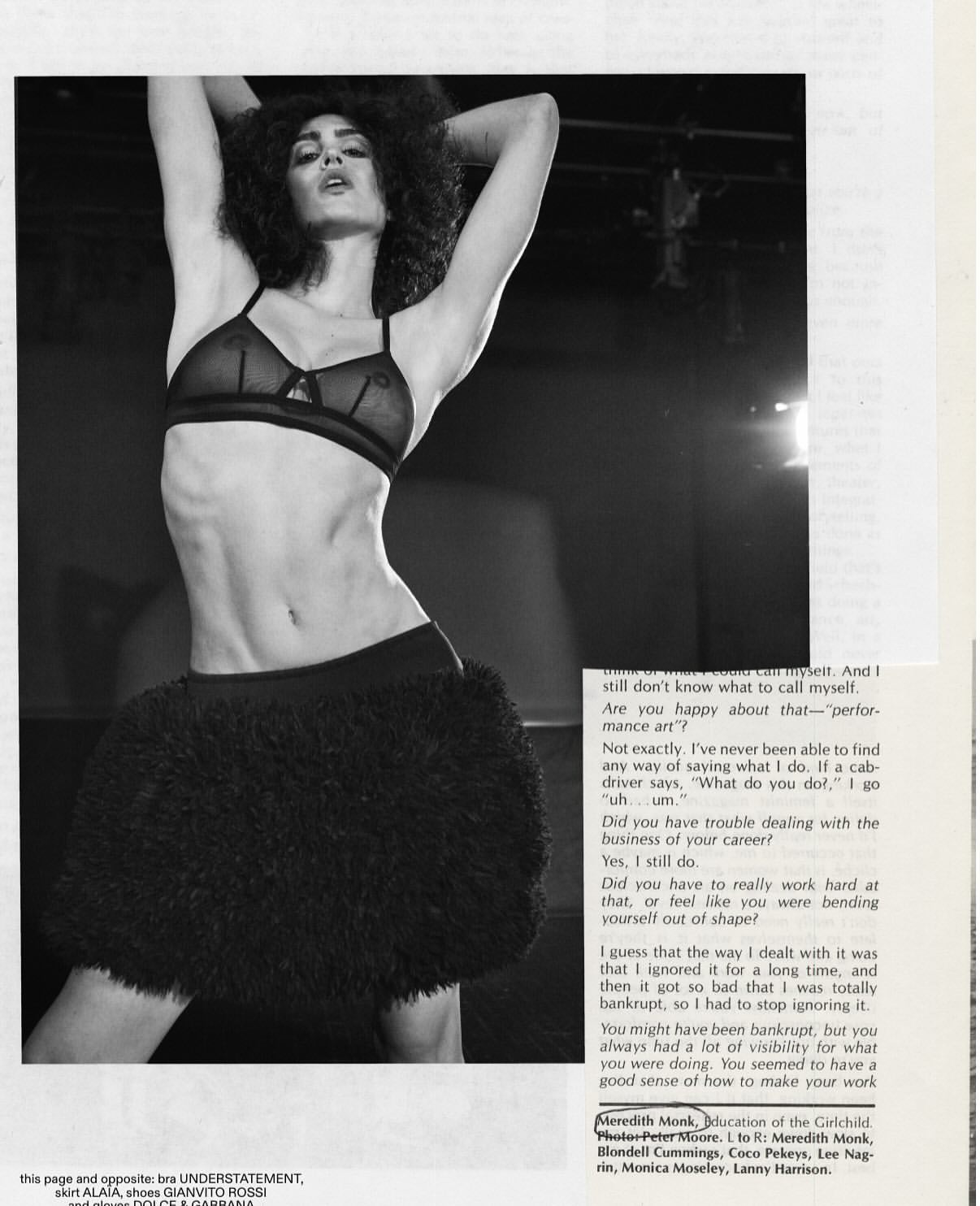 Fashion editorial shot in black and white features a woman in lingerie and a fur skirt, posing with her arms raised in an indoor setting.