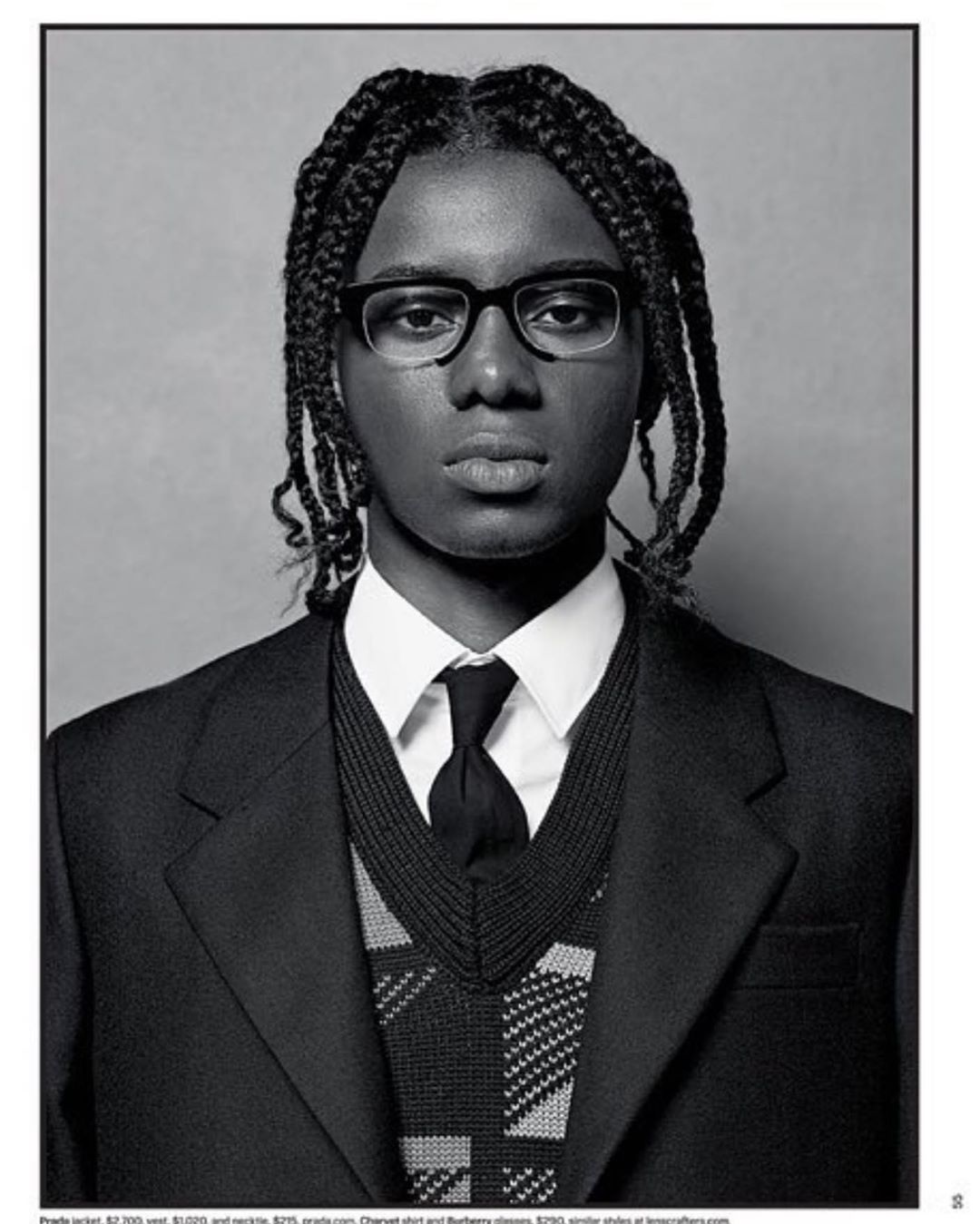 Portrait of a young adult male with braided hair, wearing glasses, a jacket, and tie, against a grey background in a studio setting.