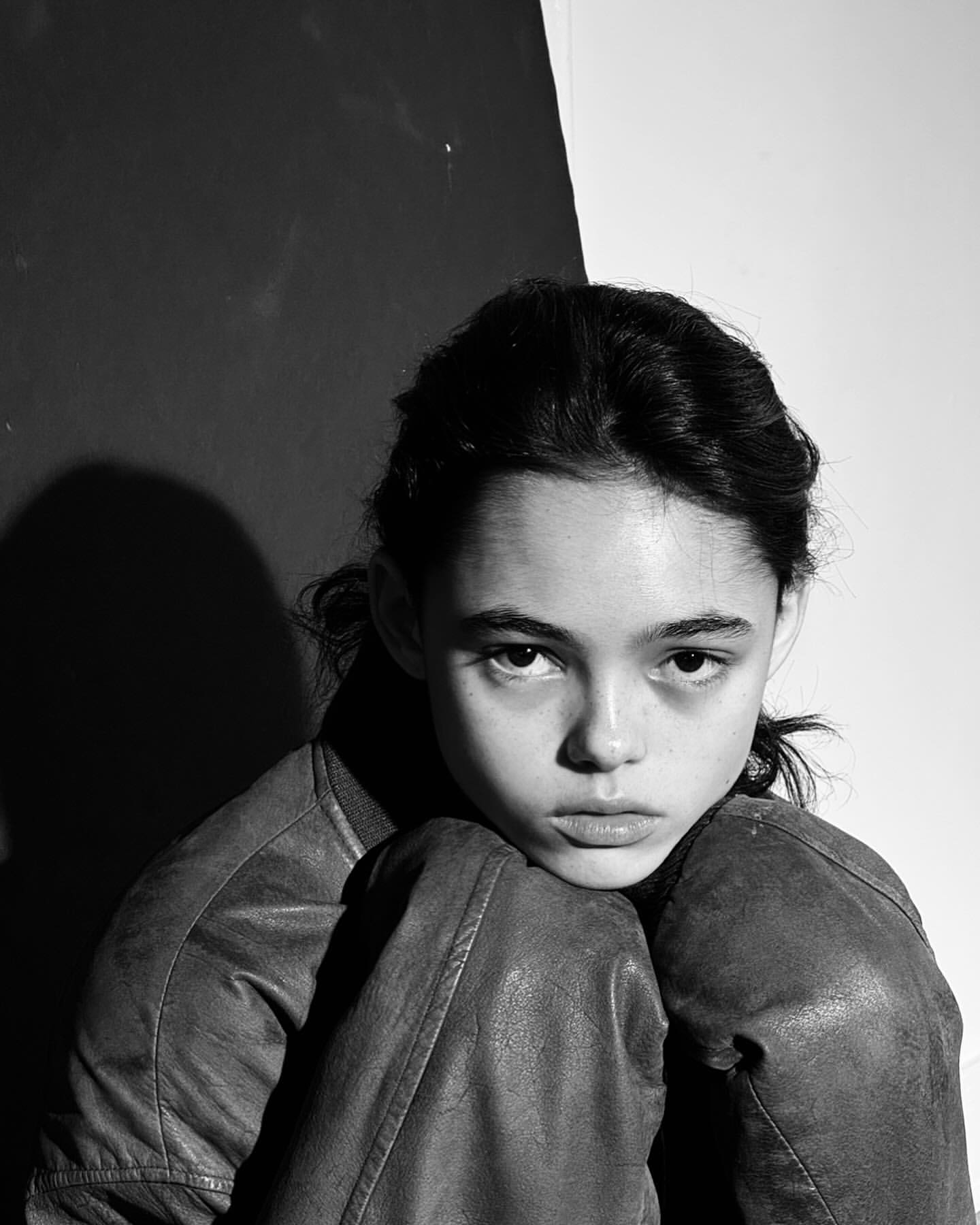 Portrait of a girl indoors in a leather jacket with dark hair, looking directly at the camera against a two-toned background.