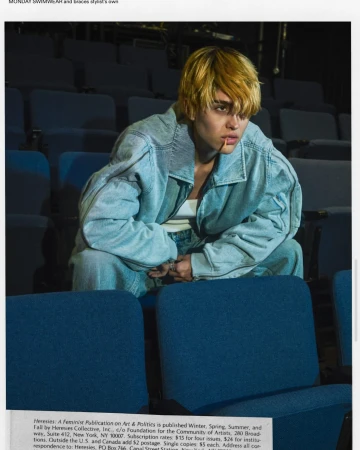 Adult in a blue denim jacket sits in a blue theatre seat, facing forward, with a toothpick in mouth and muted yellow hair.