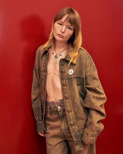 Adult wearing a denim jacket and jeans poses against a red backdrop in a studio environment, showcasing a casual fashion style.