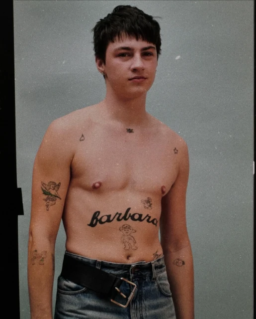 A shirtless young adult male poses in a studio, showcasing dark hair and various tattoos, including the name 'barbara' across his stomach.