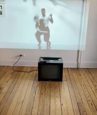 A vintage television sits in a room, displaying a video of a couple dancing on its screen, against a wall and light wood floor.