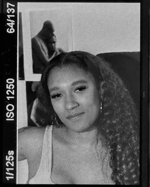 Monochrome portrait of a woman with curly hair in front of a wall decoration, captured with film grain texture and vintage aesthetic.