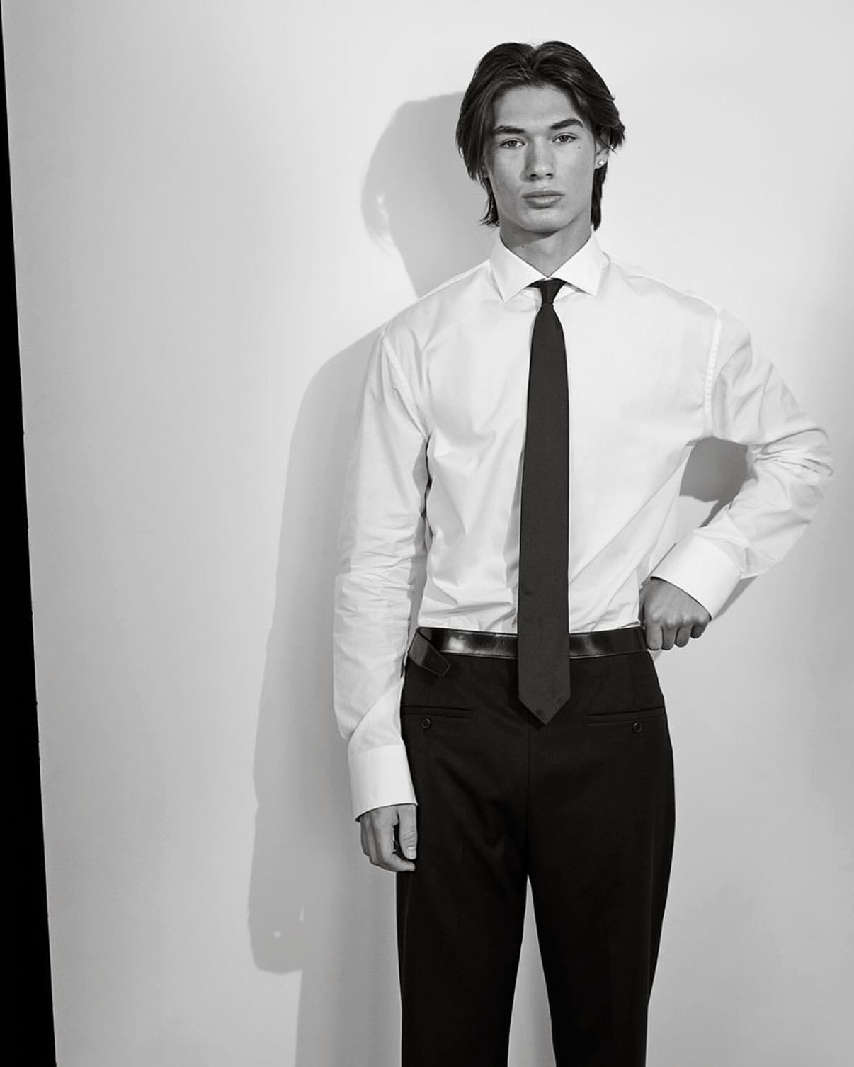 Portrait of a young man in a dress shirt and tie. Black and white studio shot with the subject standing against a white wall.