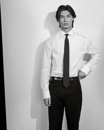 Portrait of a young man in a dress shirt and tie. Black and white studio shot with the subject standing against a white wall.
