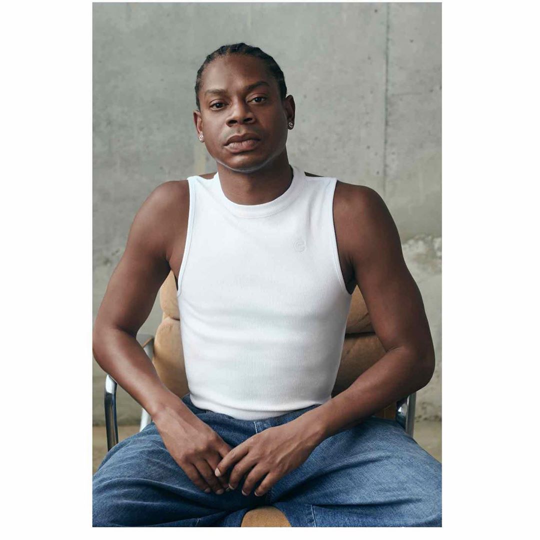 Close-up portrait of a black male in a white tank top and blue jeans sitting in a tan chair against a concrete background.