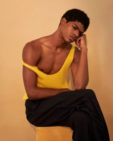 A young Black man wearing a yellow tank top and dark trousers sitting against a warm beige background, photographed by Collier Schorr.