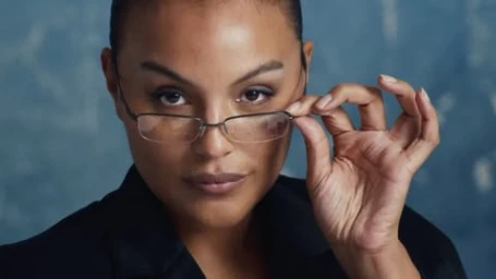 Close-up portrait of a woman adjusting her thin-rimmed glasses against a mottled blue background, directed by Columbine Goldsmith.