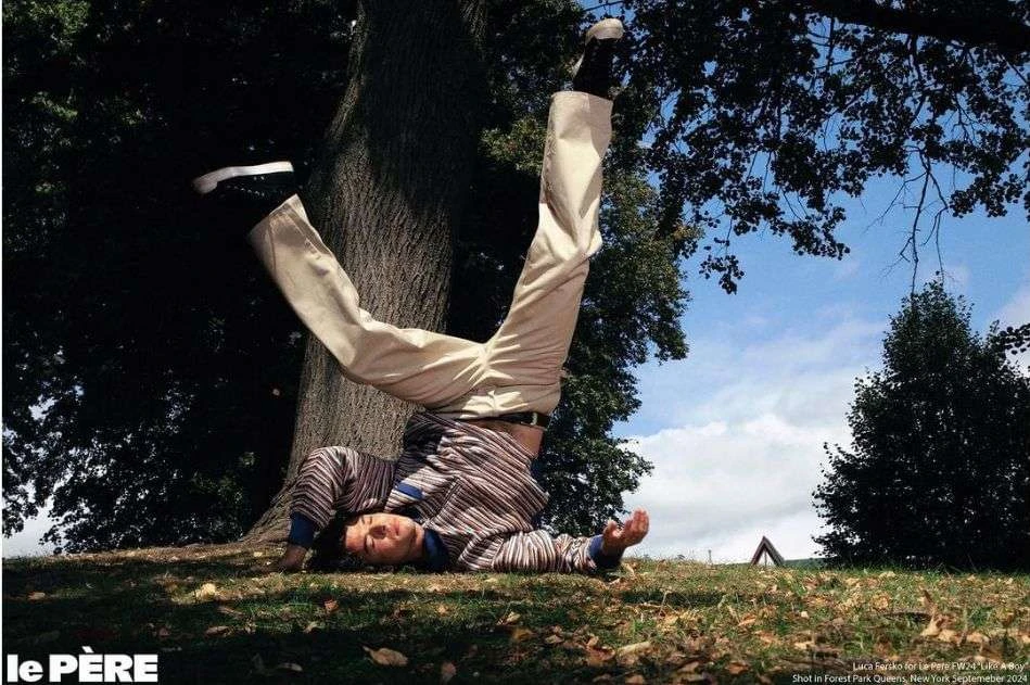 Young adult male lying upside down on green grass outdoors in a striped shirt and khaki pants, with trees and a blue sky in the background.