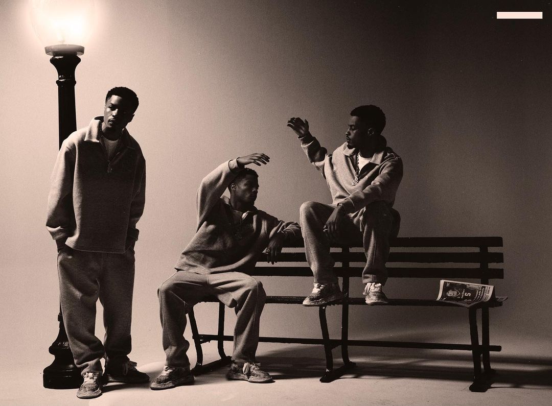 Three adult males pose on a bench outdoors next to a vintage street lamp in a sepia toned studio shot, all in matching outfits.