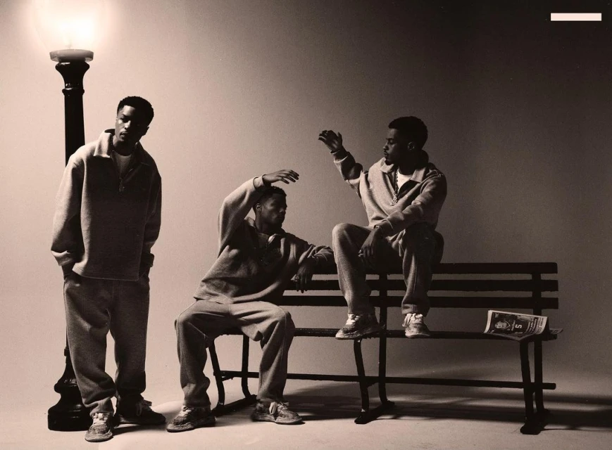 Three adult males pose on a bench outdoors next to a vintage street lamp in a sepia toned studio shot, all in matching outfits.